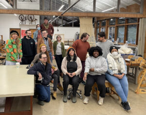Thirteen people pose casually, for a group photo in an art studio with exposed wooden beams. 