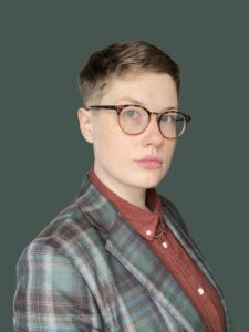 Nonbinary-presenting person with white skin, short light brown hair, and tortoiseshell glasses. They are wearing a green and brown check blazer and pumpkin-colored button-up shirt. They look into the camera with a serious and quizzical expression. 