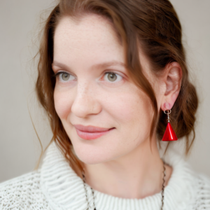Headshot of light-skinned femme with red hair, heart-shaped hairline and gray-green eyes looking off-camera (to the left), with an out of focus greige background. The femme wears a bright red cone-shaped earring, white hand-knitted crew-neck sweater and nondescript chains.
