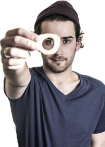 A male-presenting figure holds a small, roughly, doughnut shaped object up to the camera. Their right eye is visible through the object. The figure has light skin, dark hair, and brown eyes. They wear a dark gray tshirt and a brown beanie hat.