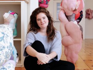 A white skinned femme with dark brown curly hair sits on the floor with her arms wrapped around one knee. She is surrounded by textile sculptures. 